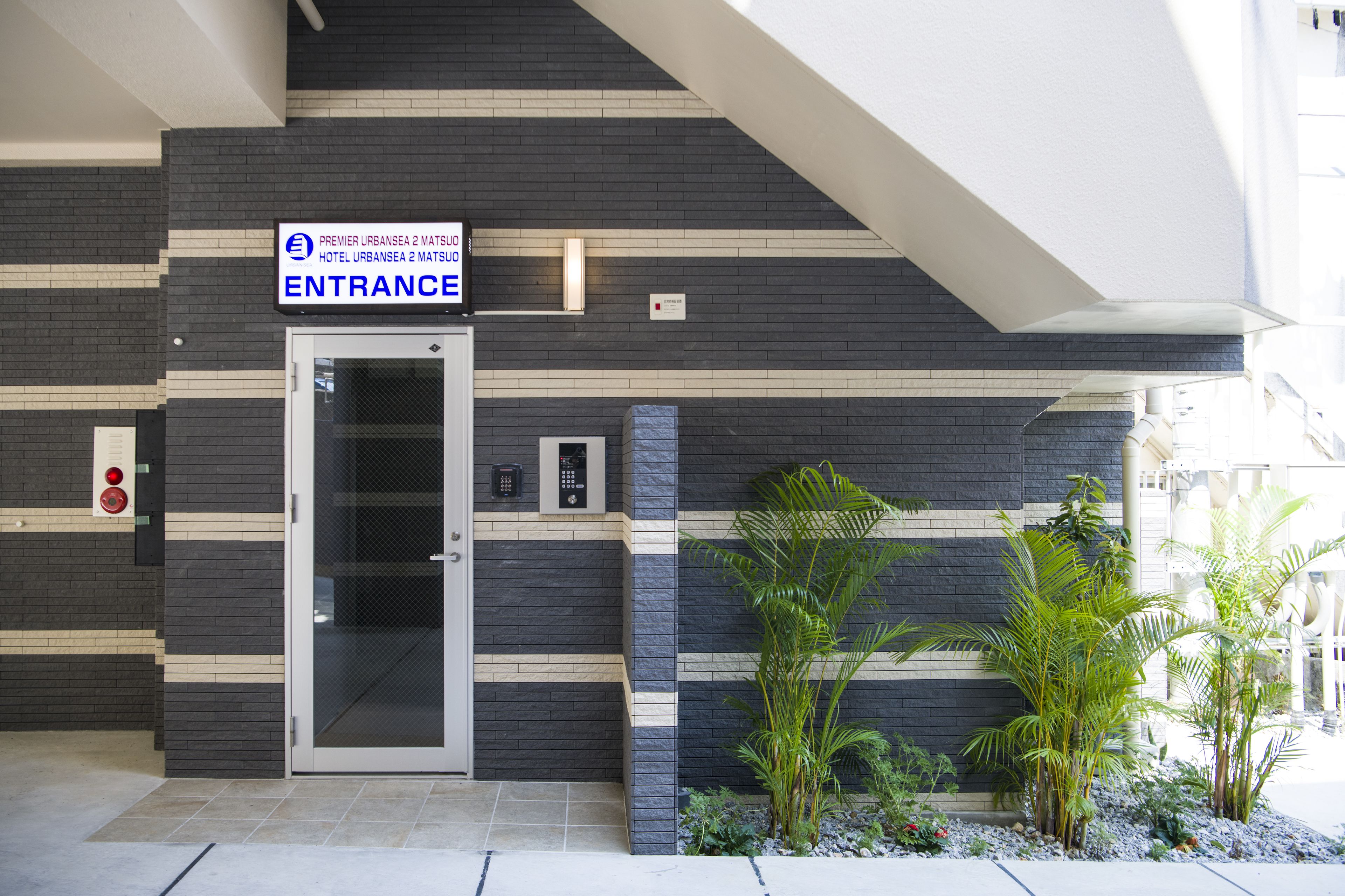 An entrance to Premier UrBansea 2 Matsudo Hotel with a modern design, featuring a glass door, a signboard, and a small garden area with plants.