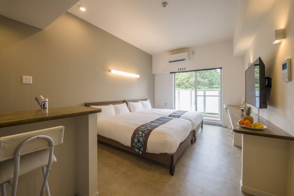 Une chambre d’hôtel avec un lit, un bureau, une chaise, une petite table avec des fruits et une fenêtre donnant sur un paysage verdoyant.