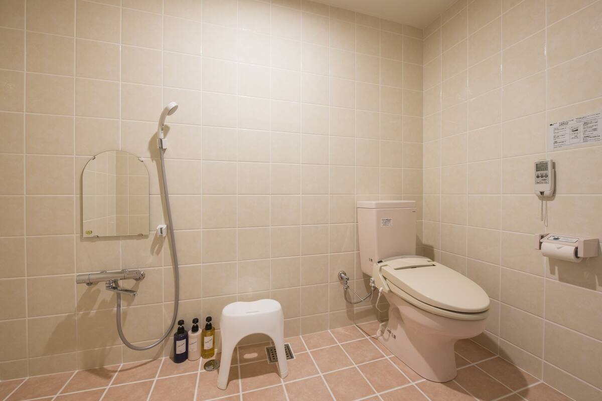 Une salle de bain avec des toilettes, une douche et un petit tabouret. On peut voir des bouteilles de produits de toilette sur le sol.