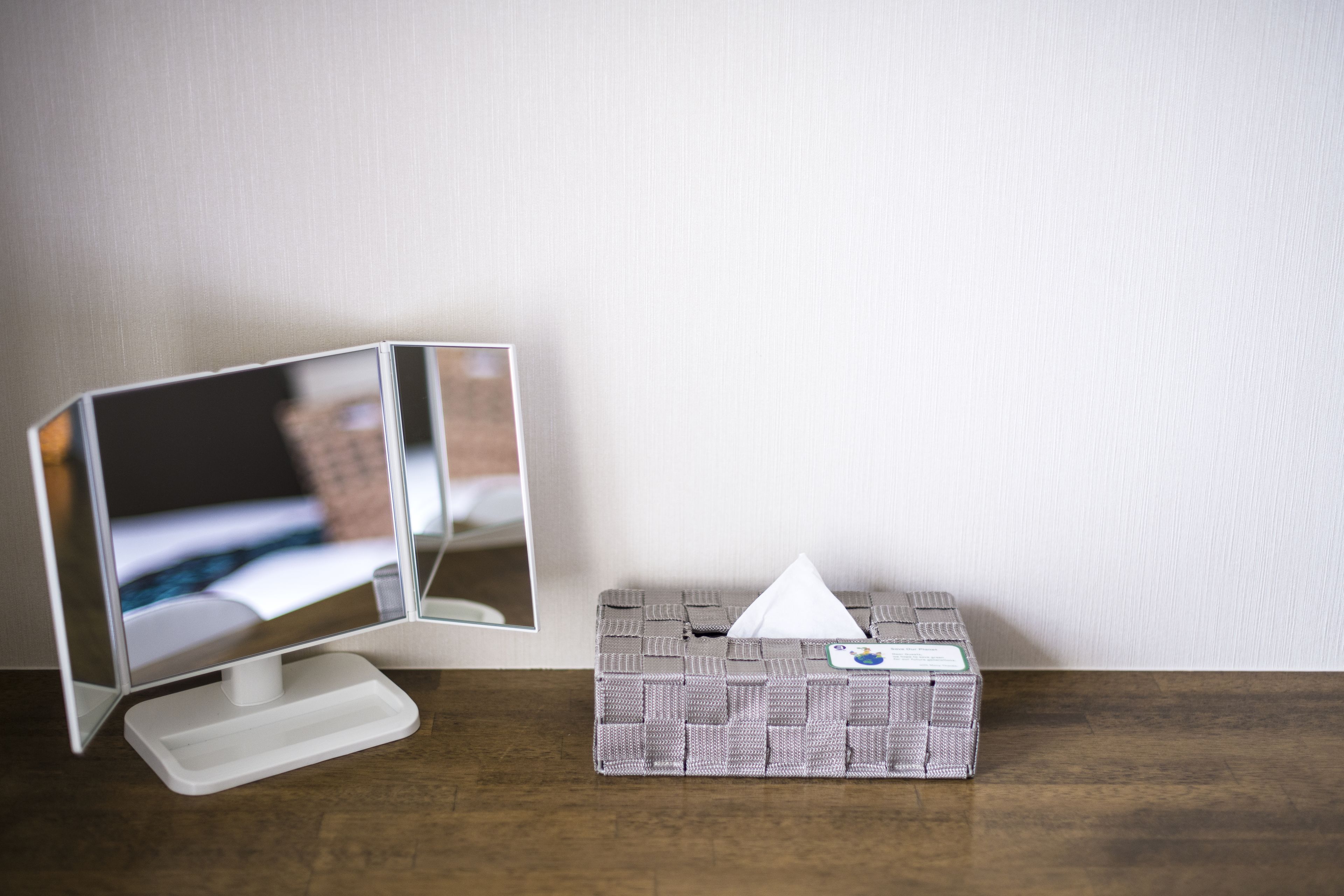 A bedside table with a bedside lamp, a tissue box, and a mirror reflecting a bed.