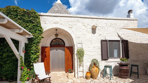 Une cour avec un bâtiment en pierre blanche, une porte en bois, des plantes en pot et une chaise blanche.