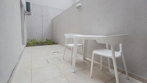 Um pátio pequeno e fechado com uma mesa e cadeiras brancas, uma planta pequena e uma parede branca.