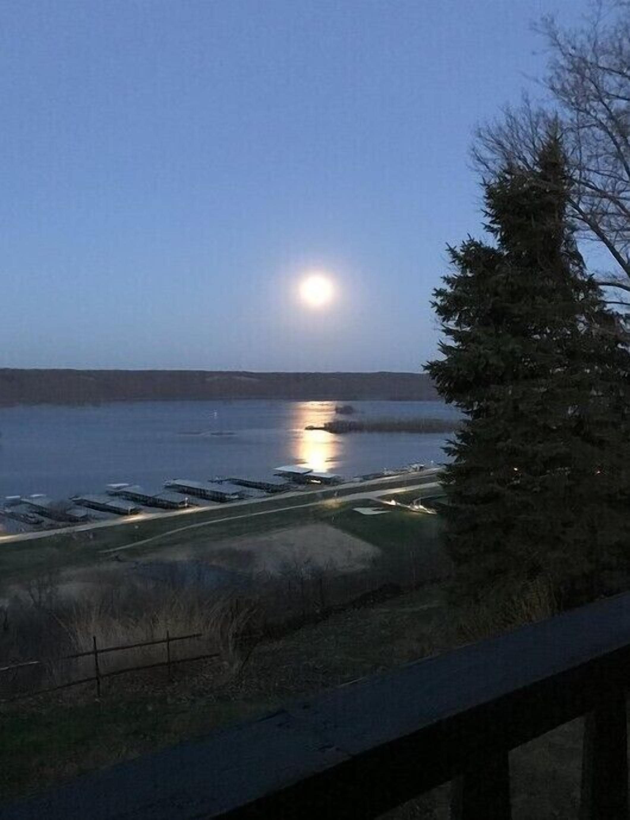 A view of a lake at dusk with a full moon reflecting on the water, surrounded by a dock and a row of buildings.