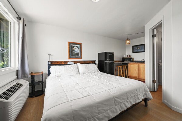 A bedroom with a bed, a bedside table, a radiator, a window with curtains, a framed picture on the wall, a kitchen area with a refrigerator, and a hanging light fixture.