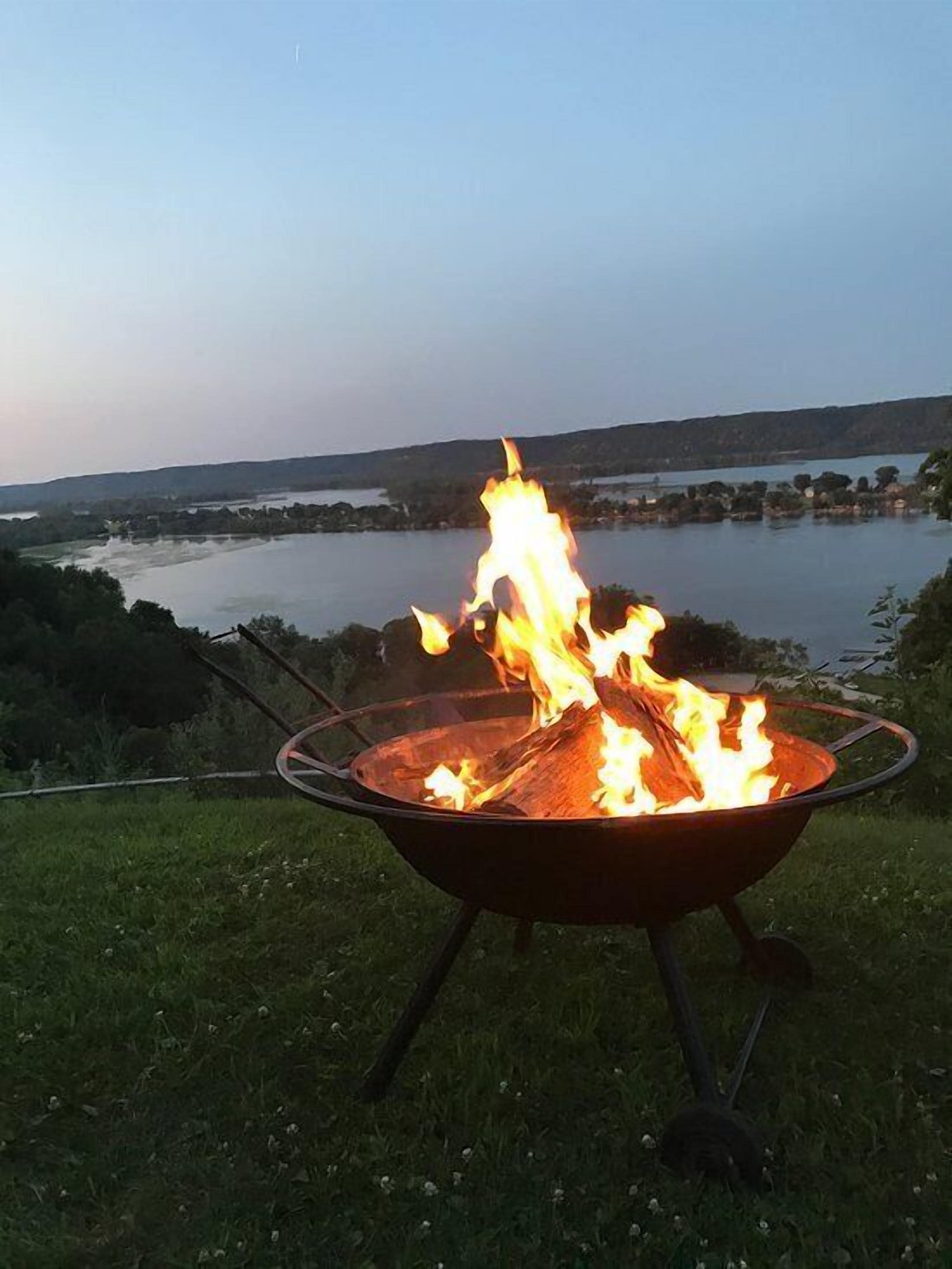 Un fuoco di campo in un braciere metallico, con un lago e delle colline sullo sfondo.