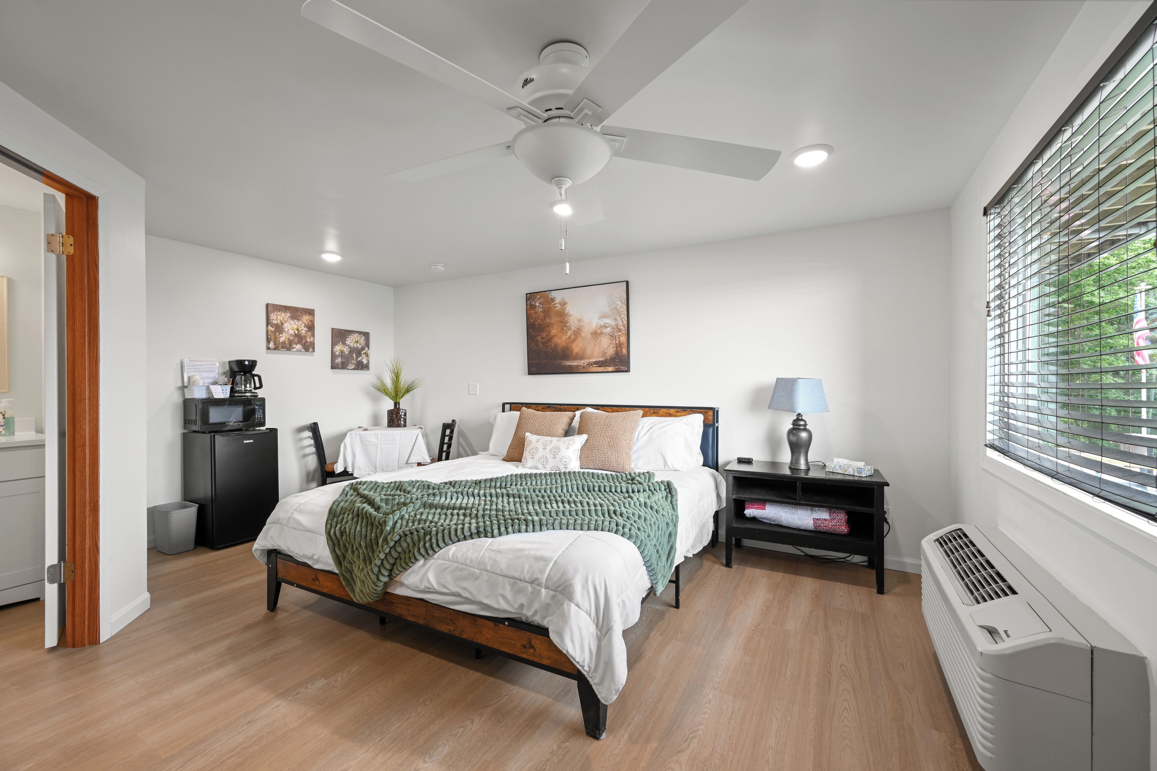 A modern bedroom with a bed, bedside table, lamp, and a view of the outdoors through a window.