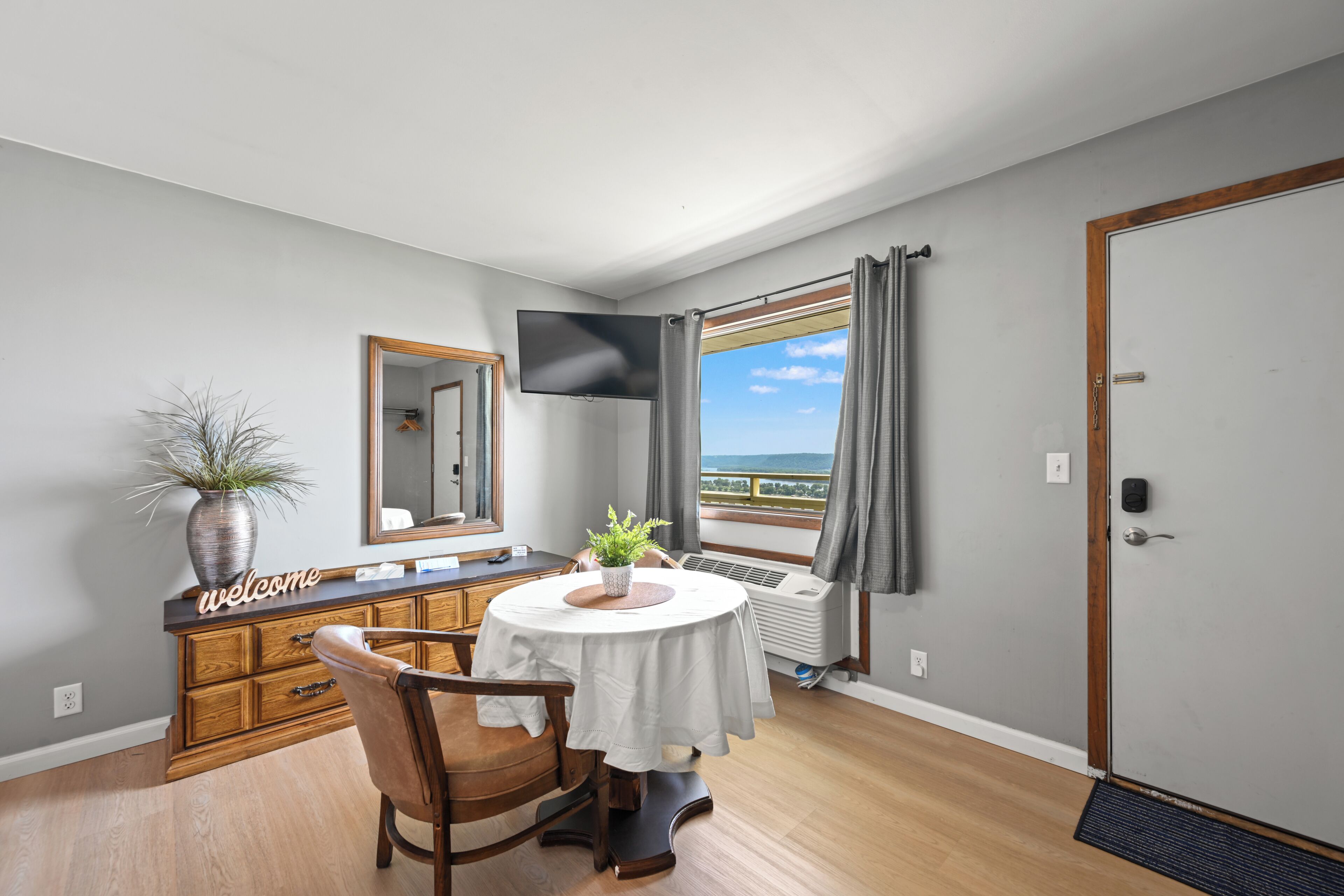 Quarto de hotel com uma mesa redonda, uma cadeira, um vaso de plantas, um espelho, uma televisão, uma janela com cortinas e uma porta.