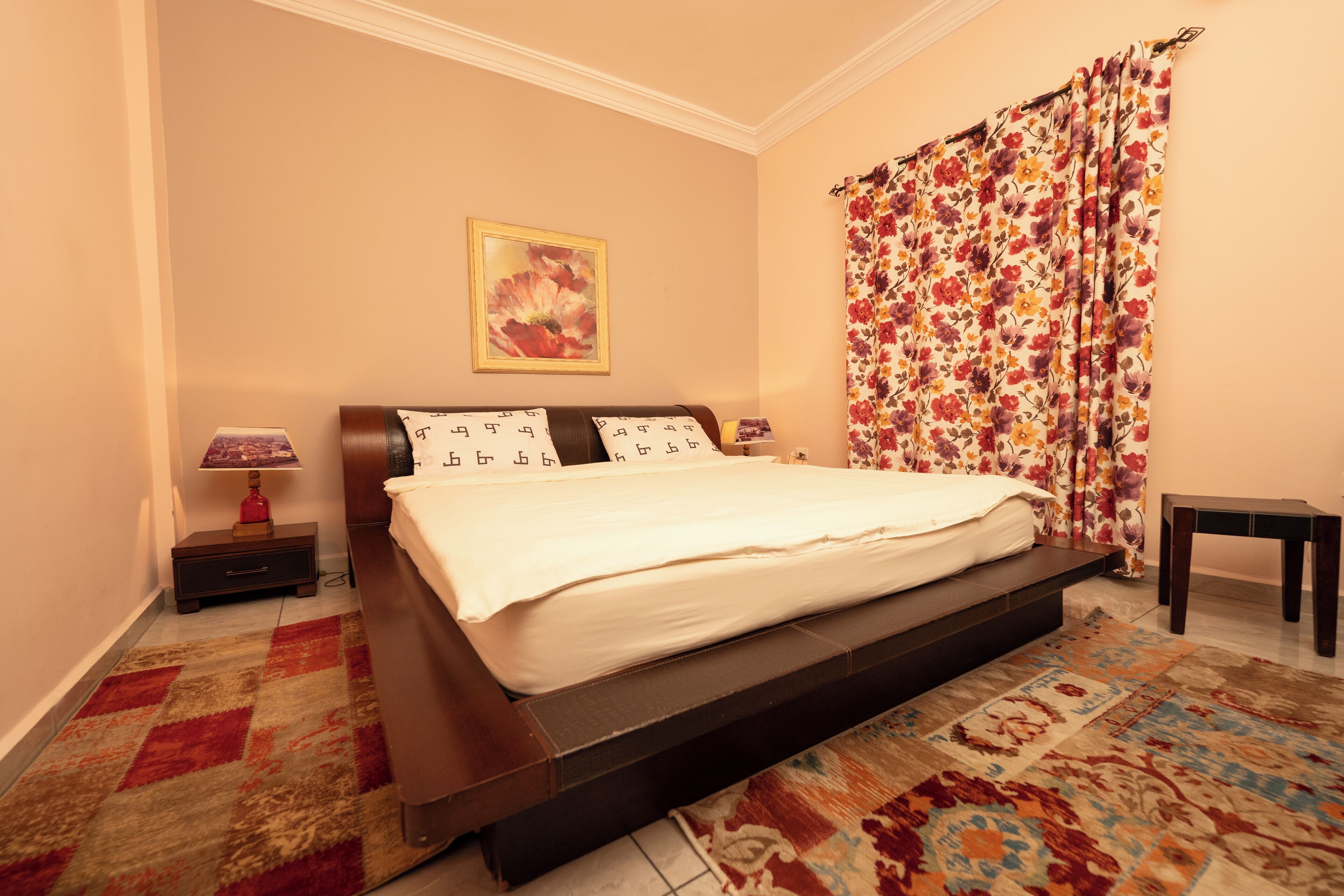 Un dormitorio con cama, mesita de noche, lámpara, un cuadro en la pared y cortina estampada.