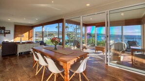A spacious dining area with a wooden table and white chairs, a sofa, and a view of the ocean through large glass doors. - Beach Side Bay Of Fires (Binalong Bay)