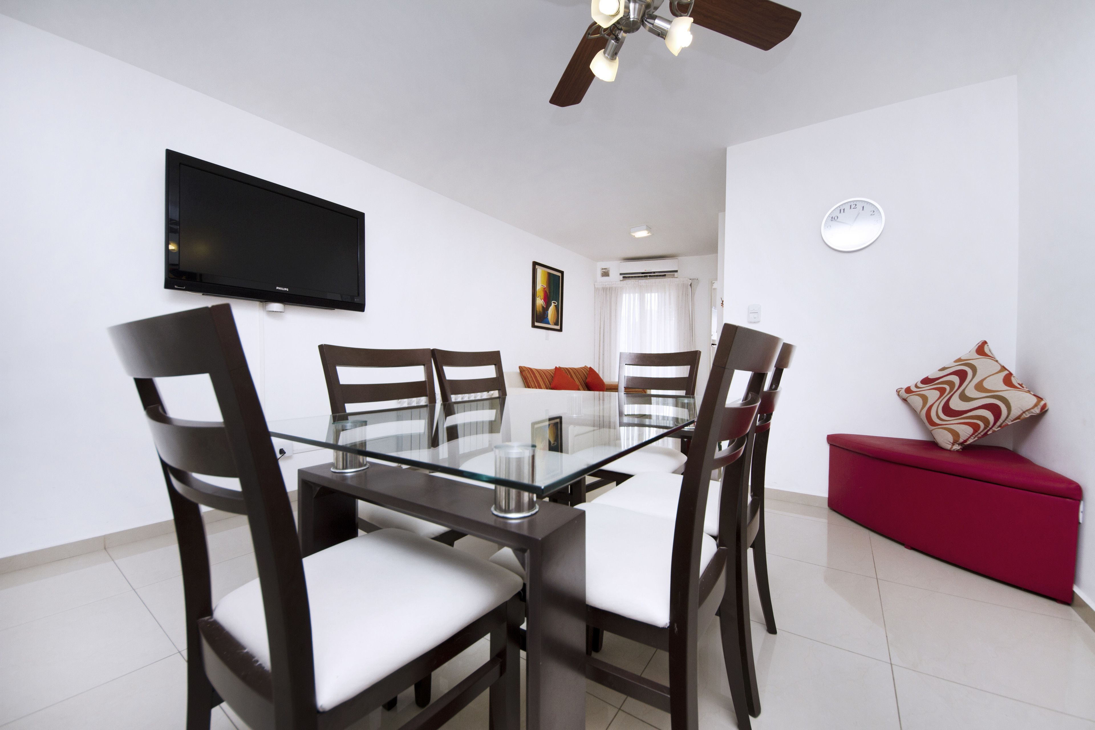 A modern living room with a glass dining table, dark wooden chairs, a red sofa, a flat-screen TV, a ceiling fan, and a wall clock.