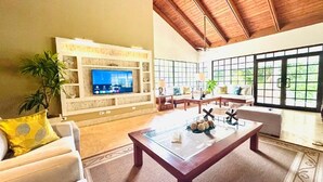 A spacious living room with a wooden ceiling, a television on a white entertainment unit, and large windows offering a view of greenery.