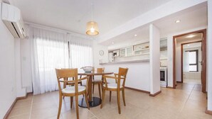 A dining area with a round table and four chairs, a hanging light fixture, and a kitchen in the background.
