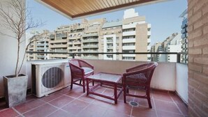 A balcony with two wicker chairs and a small table, overlooking a cityscape with multiple apartment buildings.