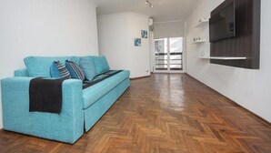 A modern living room with a teal sofa, wooden parquet flooring, a mounted TV, and a door leading to another room. - Obispo Salguero 21A (Córdoba)