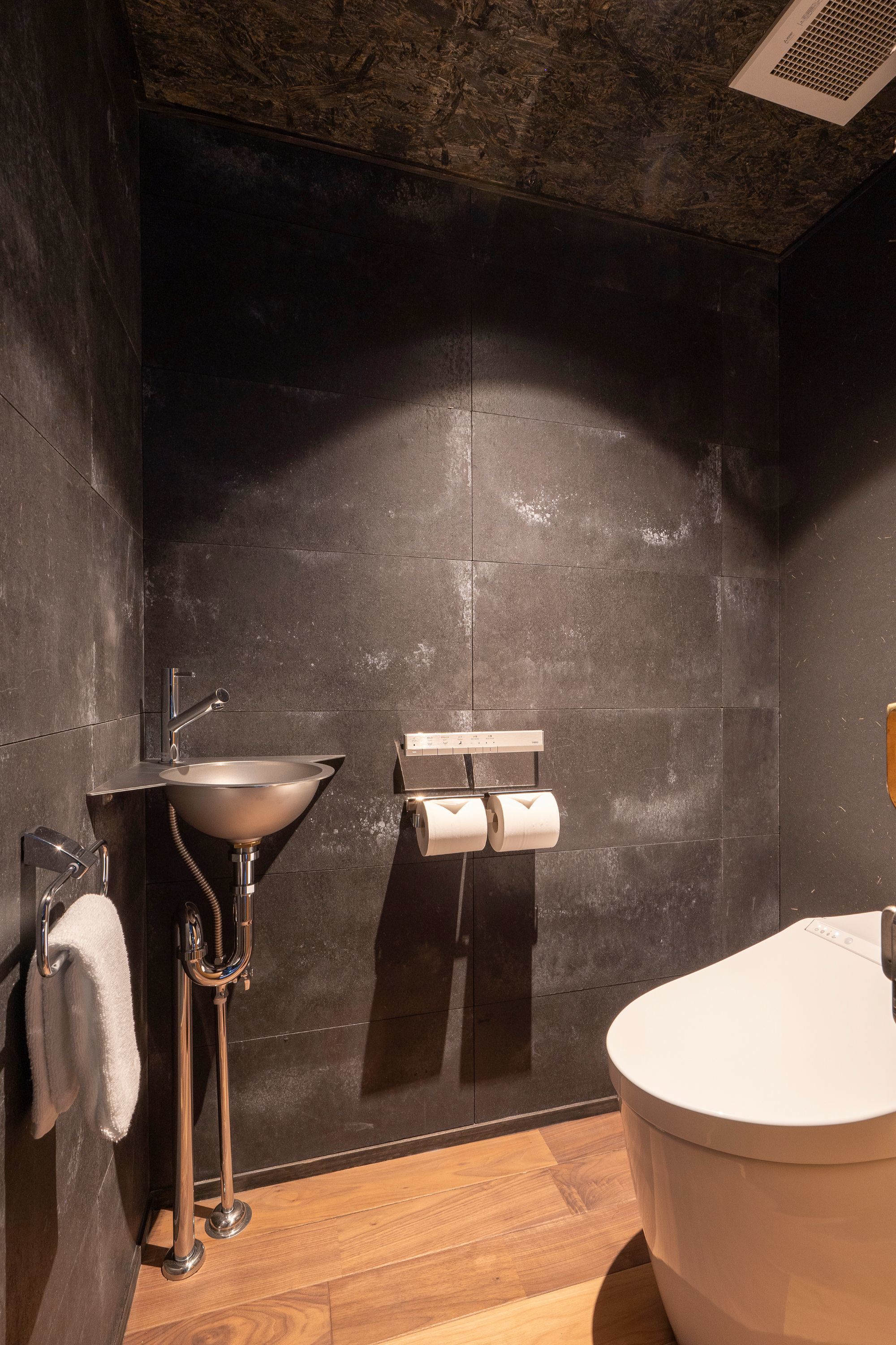 A modern bathroom with a dark tiled wall, a stainless steel sink, a toilet, and a wooden floor.