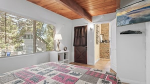 Une chambre avec un grand lit, une fenêtre avec des stores, un plafond en bois, une télévision et une porte donnant sur une autre pièce.