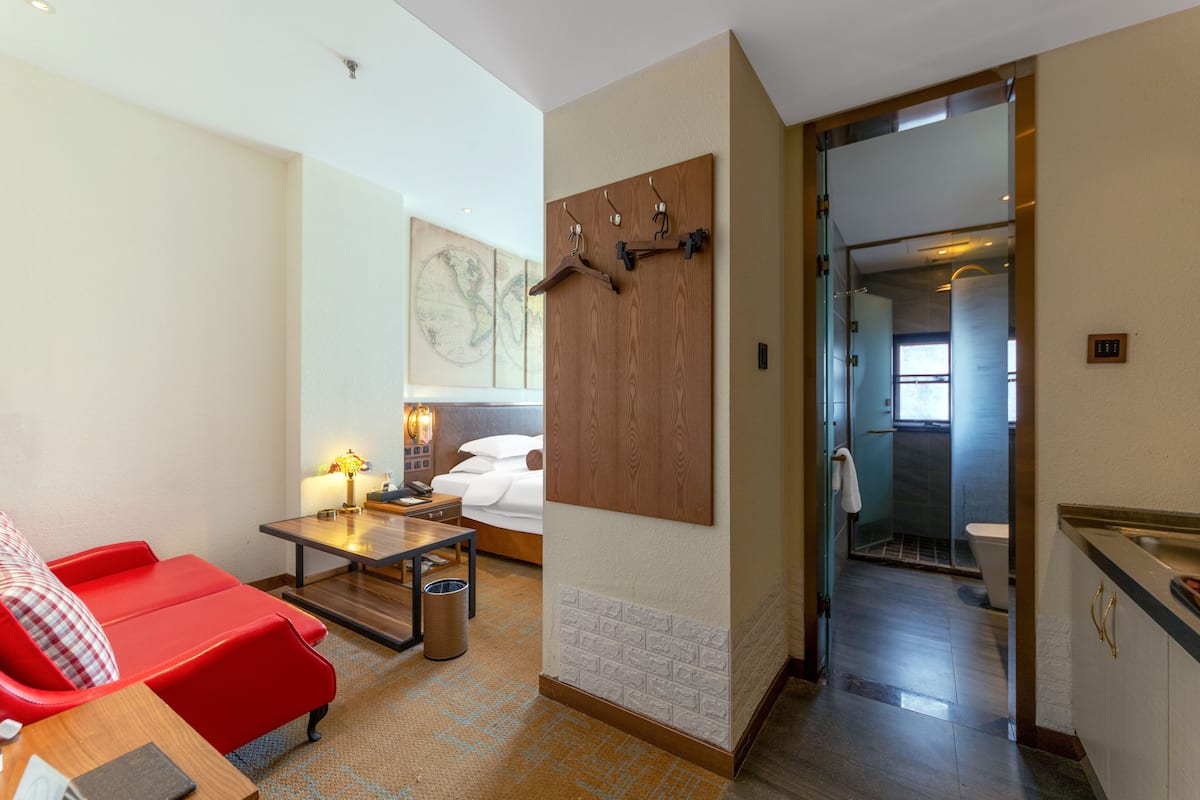 A hotel room with a red sofa, a small table with a lamp, a bed with white linens, a wooden panel wall, and a bathroom visible through an open door.