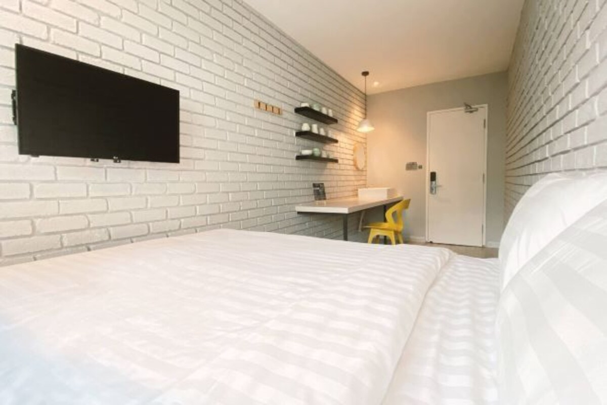 A bedroom with a white brick wall, a flat-screen TV mounted above a desk, a bed with white bedding, and a yellow chair.