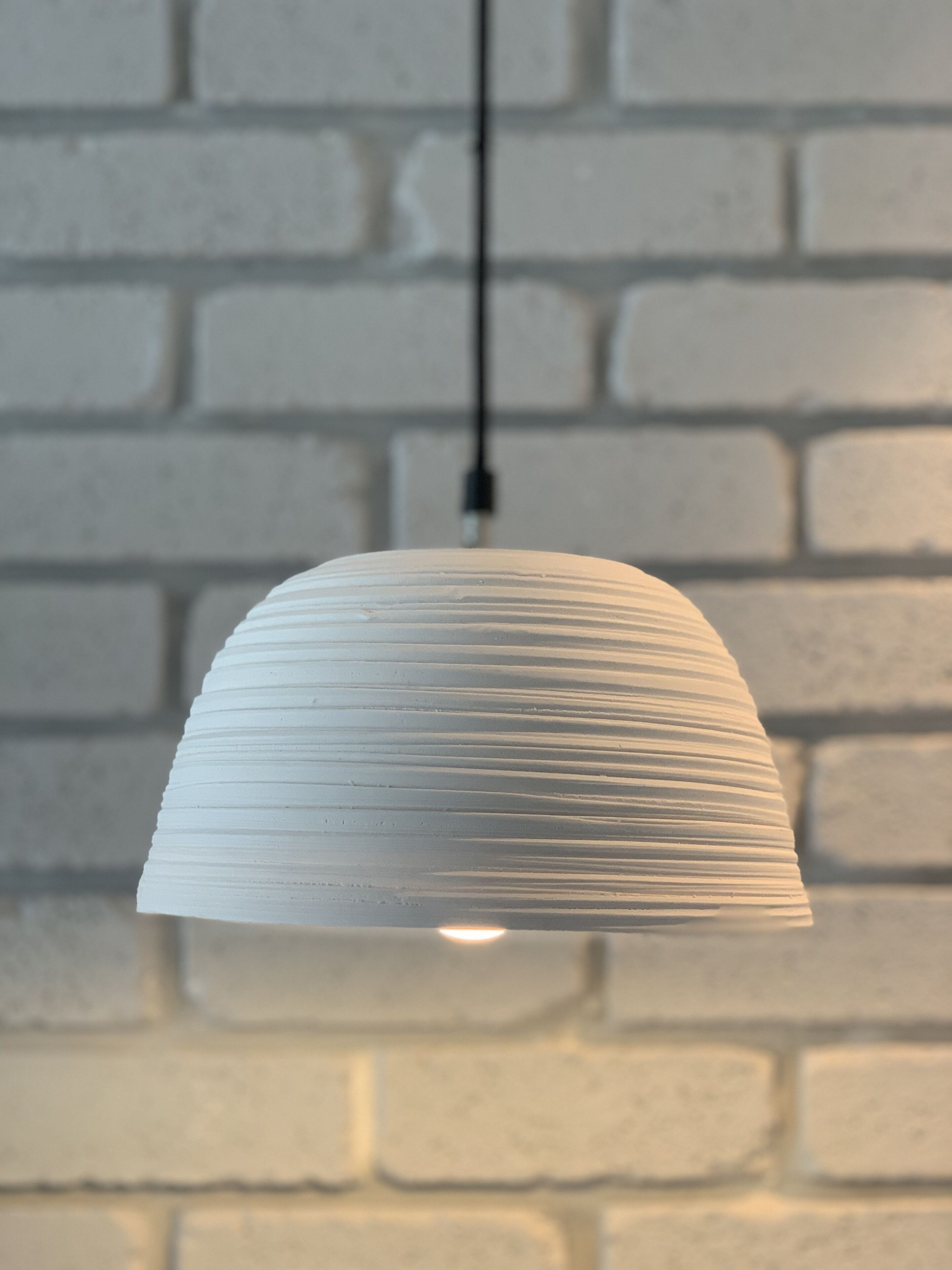 A white, ribbed pendant light hanging from a black cord against a white brick wall.