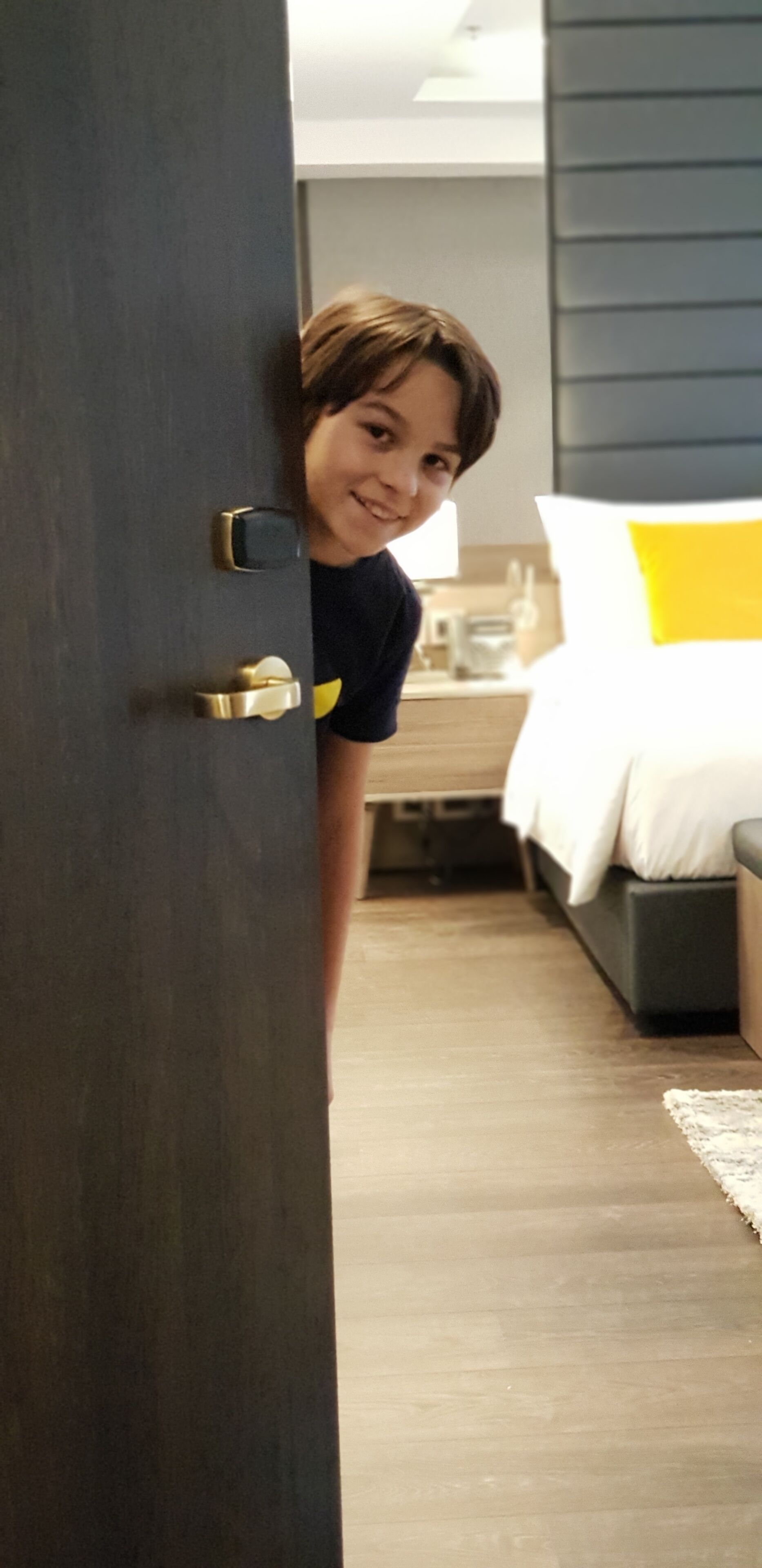 A room with a wooden floor, a bed with white bedding, and a dark-colored door with a gold handle.