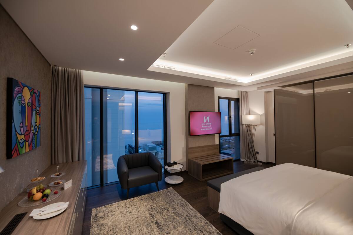 Una habitación de hotel moderna con una cama grande, un televisor de pantalla plana, una zona de estar con una silla y una mesita pequeña, un ventanal con vista a la ciudad y un cuadro en la pared.