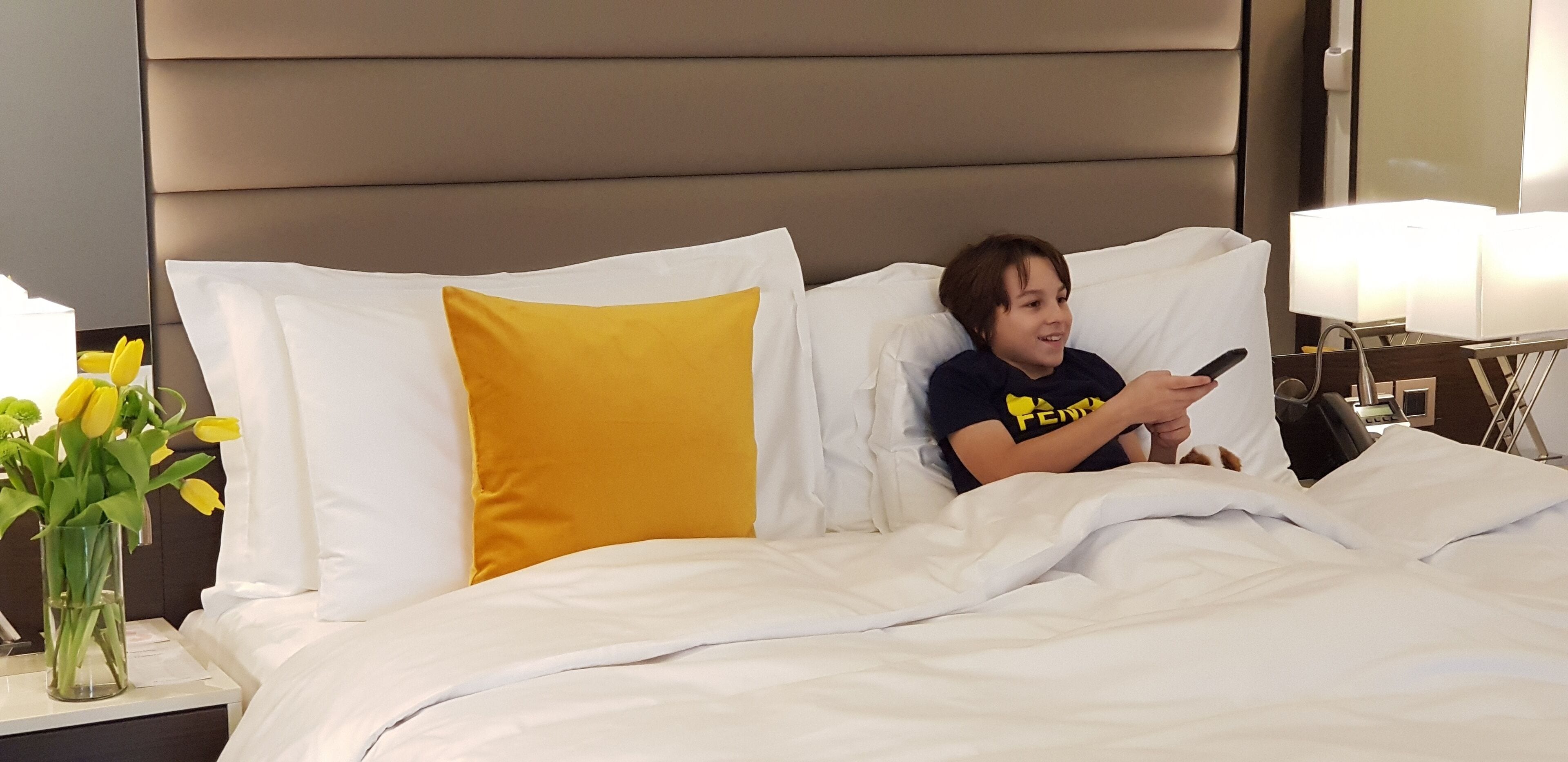 Una habitación de hotel con una cama, almohadas blancas y amarillas, una mesita de noche con un florero y una lámpara.
