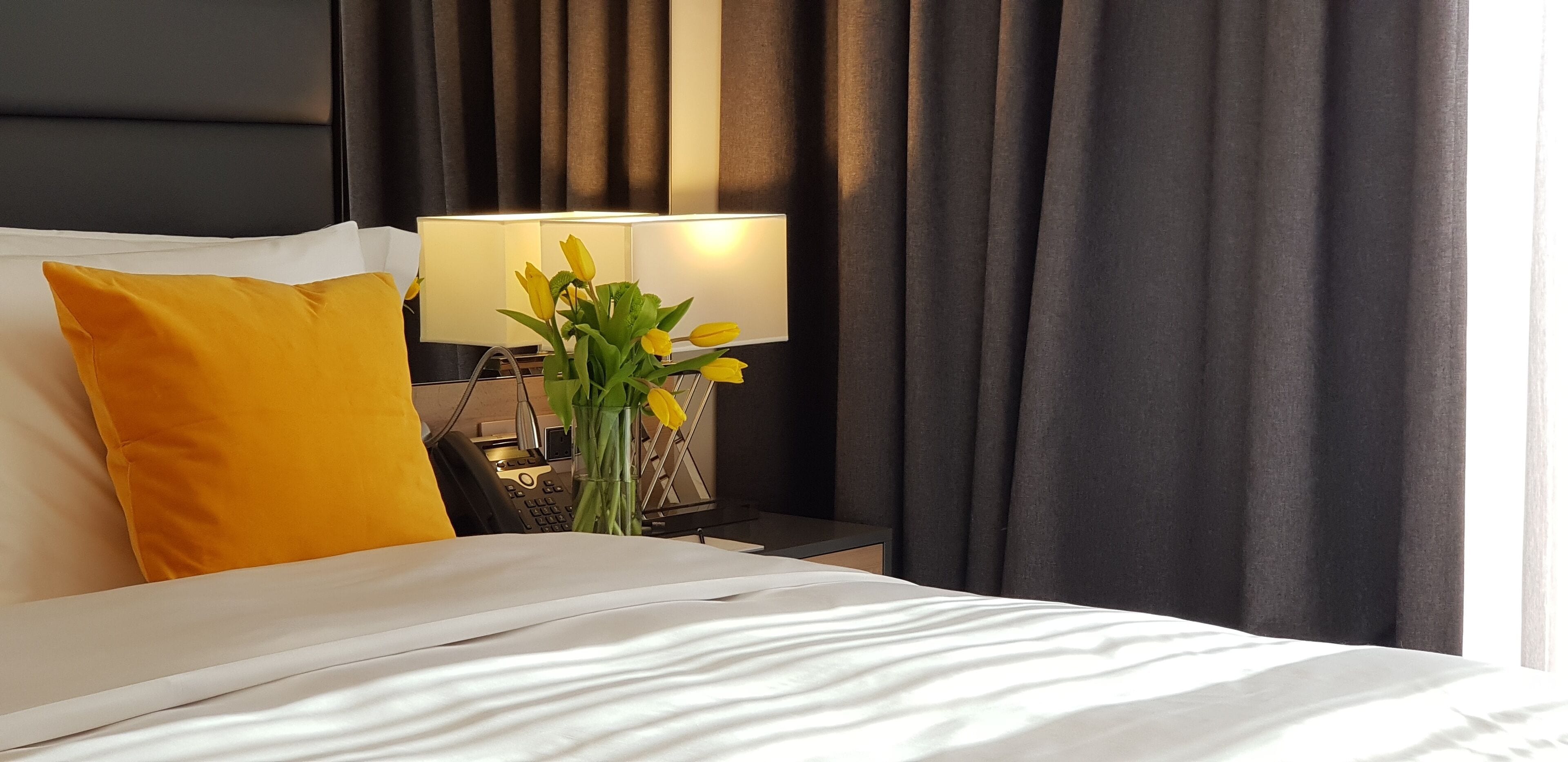 Un lit bien fait, avec du linge de lit blanc et un seul oreiller jaune, une lampe de chevet et un vase rempli de fleurs jaunes sur une table de chevet.