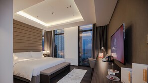 Una habitación de hotel moderna con una cama grande, un escritorio con televisor de pantalla plana, una zona de estar con vistas a la ciudad y un ventanal amplio.