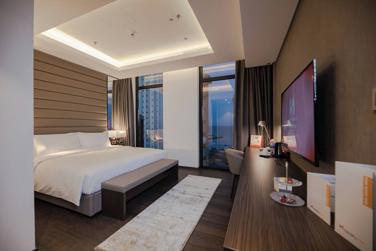 Una habitación de hotel moderna con una cama grande, un escritorio con televisor de pantalla plana, una zona de estar con vistas a la ciudad y un ventanal amplio.