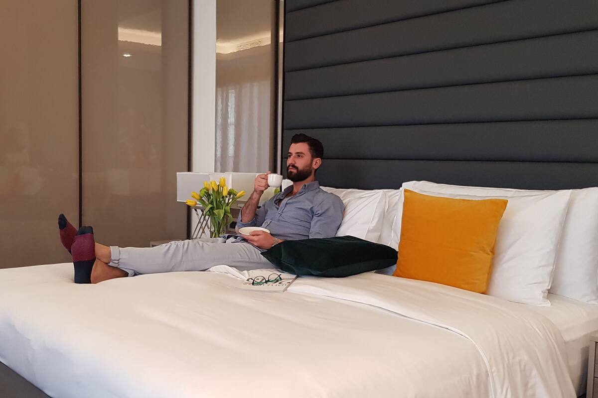 Una persona recostada en una cama en una habitación de hotel moderna, con un ventanal grande, una mesita de noche y una lámpara de noche.