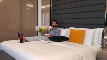 Una persona recostada en una cama en una habitación de hotel moderna, con un ventanal grande, una mesita de noche y una lámpara de noche.