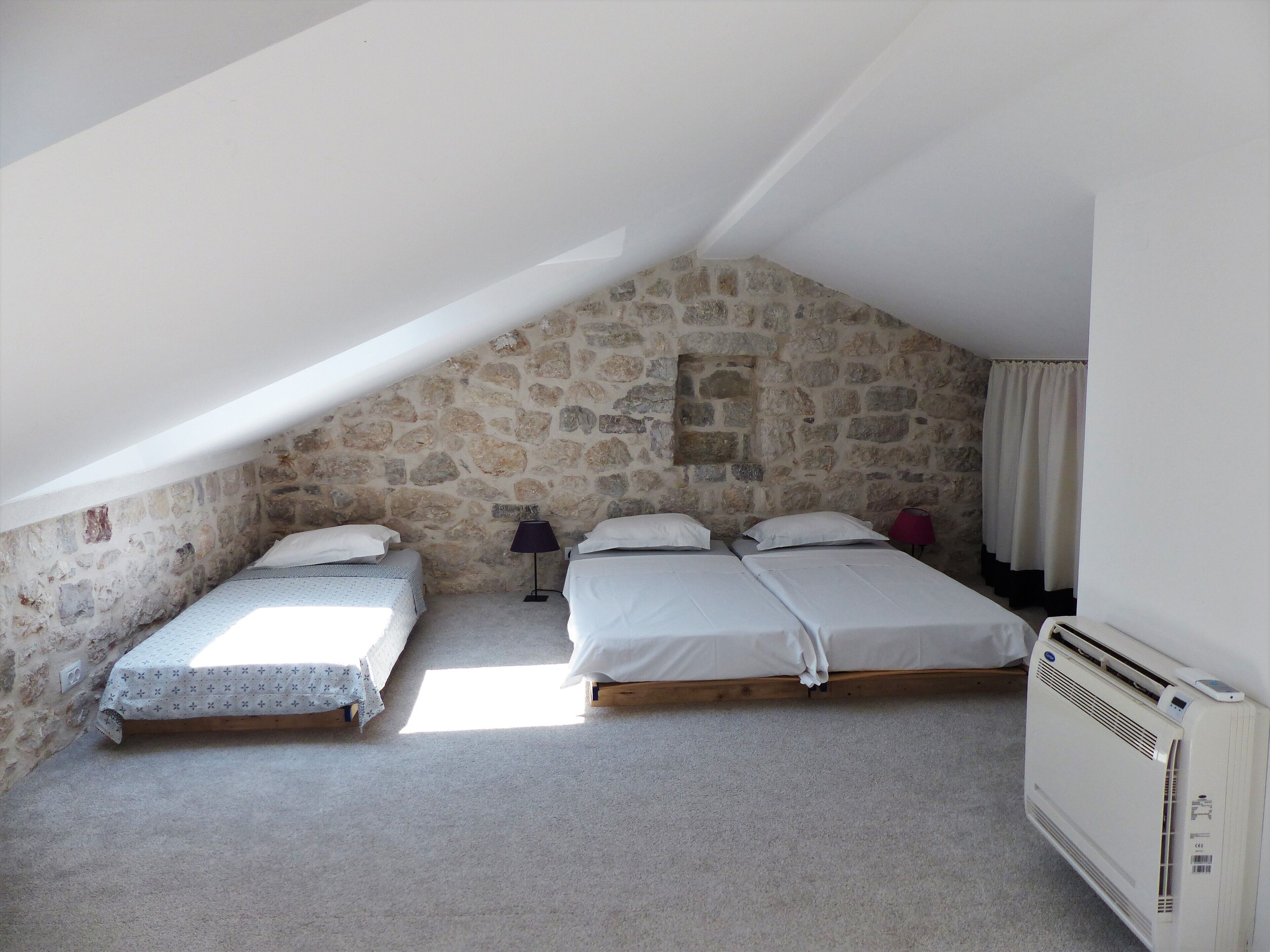 A small, stone-walled attic bedroom with two beds, a heater, and a window allowing natural light.