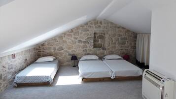 A small, stone-walled attic bedroom with two beds, a heater, and a window allowing natural light.