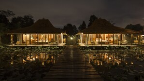 Eine friedliche Szene bei Nacht: ein Resort mit zwei Gästehäusern, die durch einen hölzernen Weg über einen Teich verbunden sind, umgeben von üppiger Vegetation.