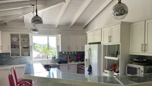 Une cuisine moderne avec des meubles blancs, un grand îlot central doté de tabourets, un réfrigérateur, un four à micro-ondes et une fenêtre donnant sur un espace verdoyant.