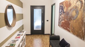 Un pasillo con piso de madera, un banco blanco con cojín negro, una estantería blanca con libros, un cuadro abstracto grande y una puerta con vidrio esmerilado.