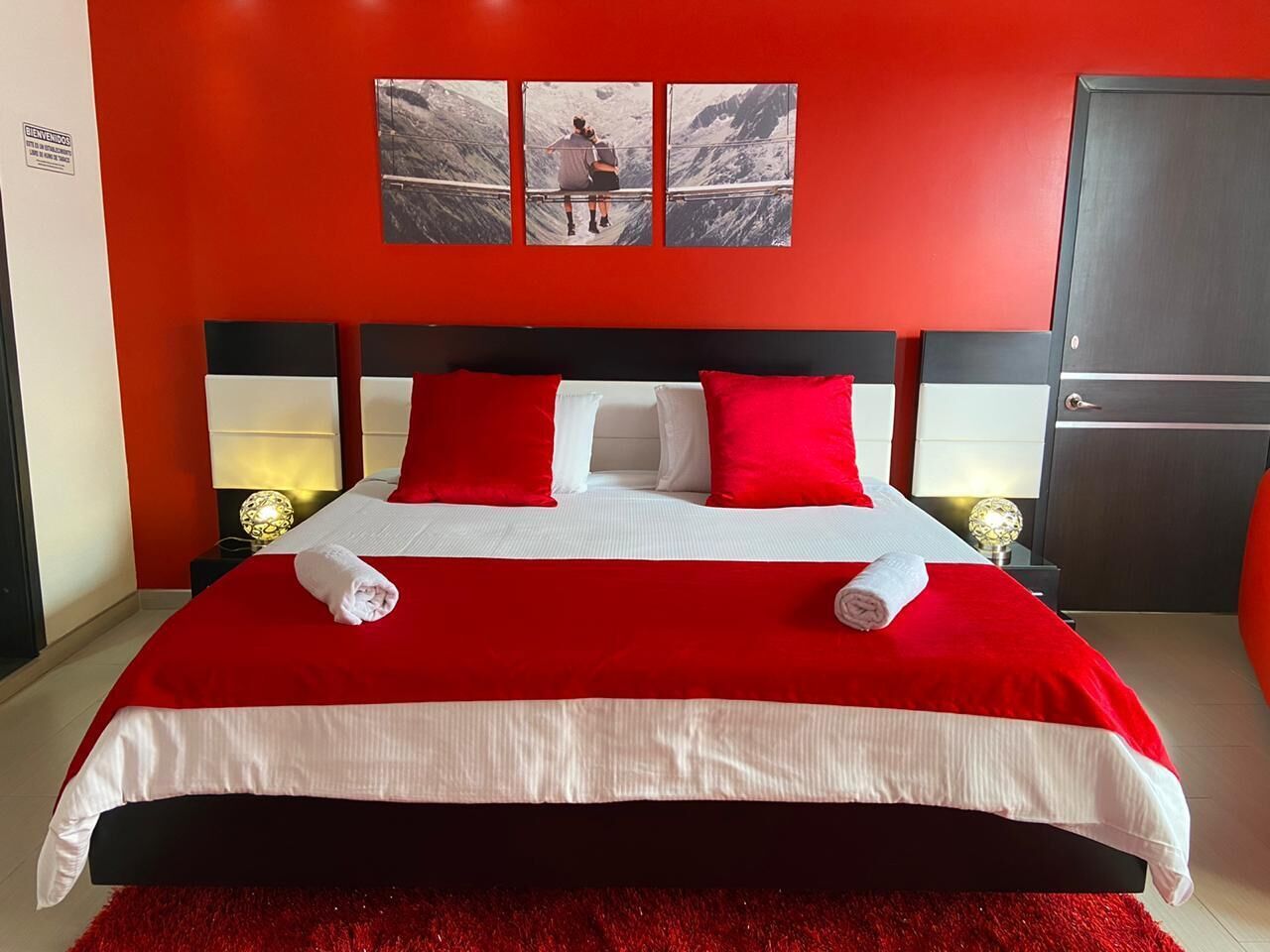 A hotel room with a large bed, red and white bedding, two bedside lamps, and a red carpet.
