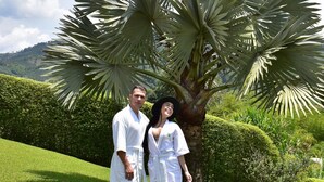 Deux personnes se tiennent dans un jardin luxuriant, avec un grand palmier et un arbuste taillé avec soin en arrière-plan.
