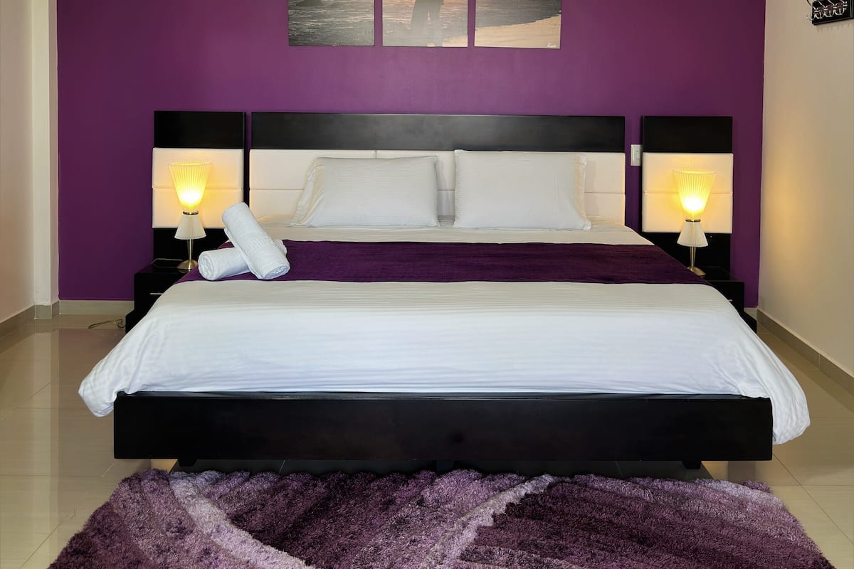 A hotel room with a large bed, two bedside tables with lamps, a purple wall, and three framed pictures above the bed.