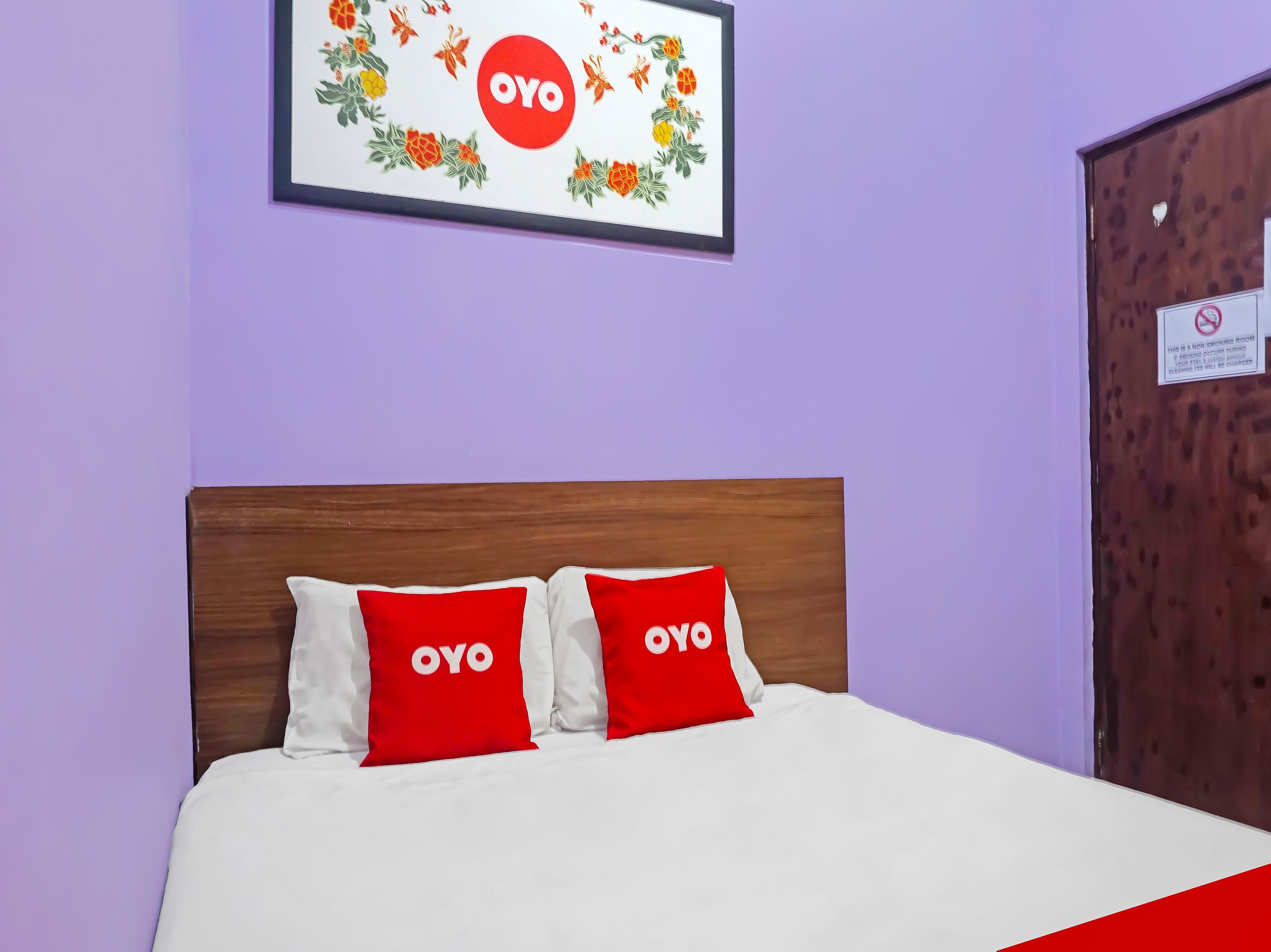 A hotel room with a bed, two pillows with the logo 'OYO', a framed picture on the wall, and a door with a no-smoking sign.