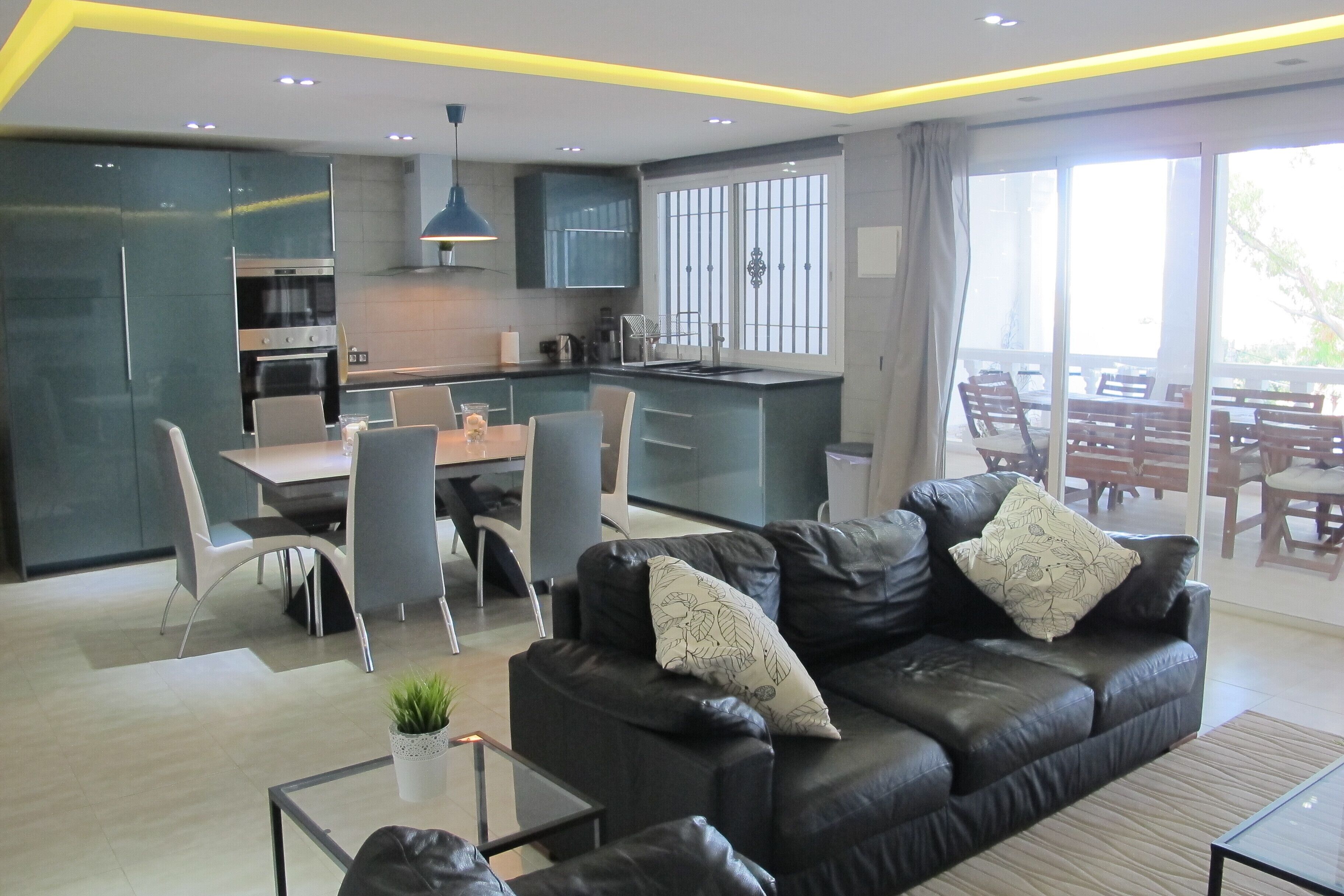Una cocina moderna con gabinetes oscuros, una zona de comedor con mesa y sillas, un sofá de cuero con cojines decorativos, y un amplio ventanal corredera que da a un patio exterior.