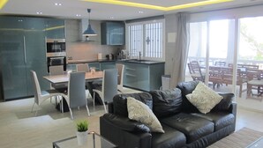 Eine moderne Küche mit dunklen Schränken, ein Essbereich mit Tisch und Stühlen, ein Ledersofa mit Zierkissen und eine große Schiebeglastür, die auf eine Außenpatio führt.