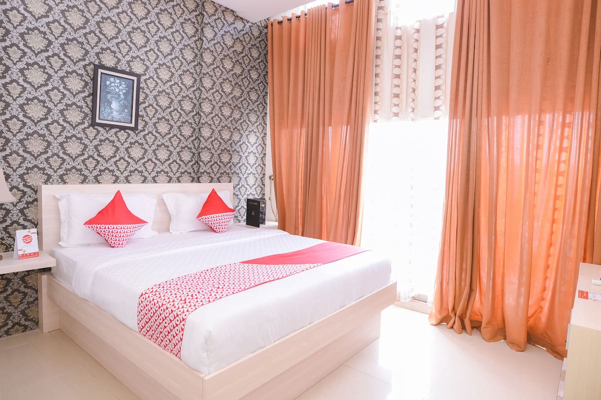 Un lit bien fait, recouvert d’une couvre-lit blanche et rouge, avec une tête de lit en bois et deux oreillers rouges. Un tableau encadré est accroché au mur au-dessus du lit, et une table de chevet avec une lampe et une boîte se trouve à côté.