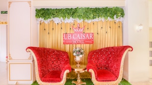 A room with two red velvet chairs, a small round table with a vase of flowers, and a backdrop with 'UB CAISAR HOTEL' signage. - Capital O 854 Ub Caisar Hotel (Banda Aceh)