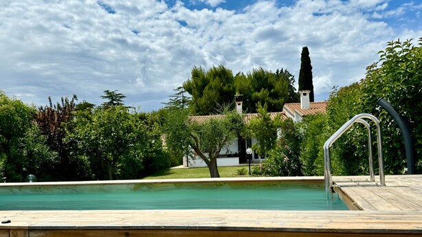 A wooden deck with a swimming pool, surrounded by greenery and a building with a tiled roof. - N084 - Numana, villa with garden and pool (Numana)
