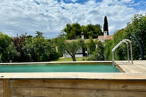 A wooden deck with a swimming pool, surrounded by greenery and a building with a tiled roof. - N084 - Numana, villa with garden and pool (Numana)