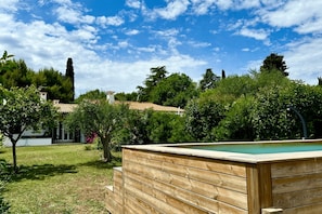A wooden deck with a pool, surrounded by greenery and a clear sky.