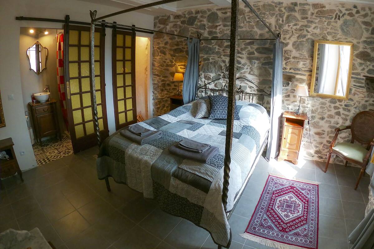 Un dormitorio con una cama con dosel, paredes de piedra, una mesita de noche de madera, un espejo y una alfombra roja con estampado.