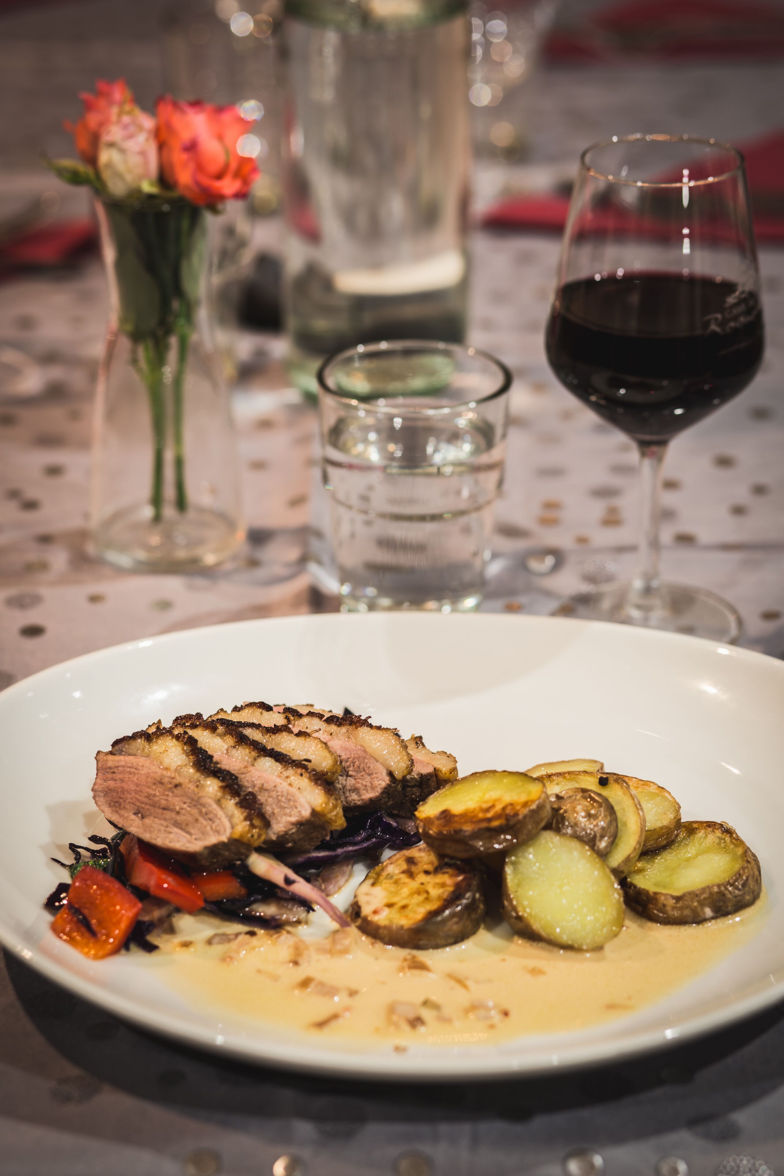 Un plato de comida con carne en rodajas, patatas asadas y verduras, acompañado de una copa de vino tinto y un florero con flores.