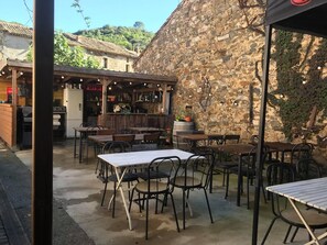 Un café en plein air avec des murs en pierre rustique, des tables et des chaises en bois, et un coin bar agrémenté d’étagères et de plantes en pot.